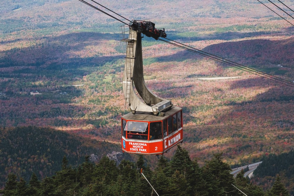 A red cable car with "Franconia Notch State Park" and "Cannon" written on it above mountains with autumn foliage.