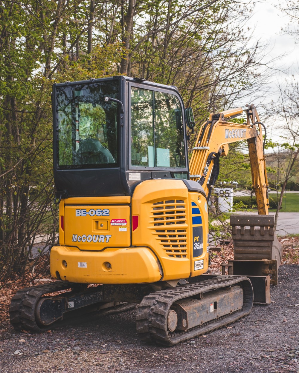 An empty yellow excavator with "McCourt" written on the arm, parked on a gravel surface with trees in the background.