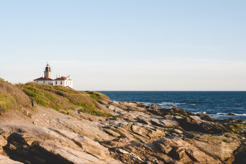 A coastal landscape featuring a lighthouse with an attached building on a cliff, overlooking a rocky shoreline with the sea in the background. There are people walking on the rocks near the water.