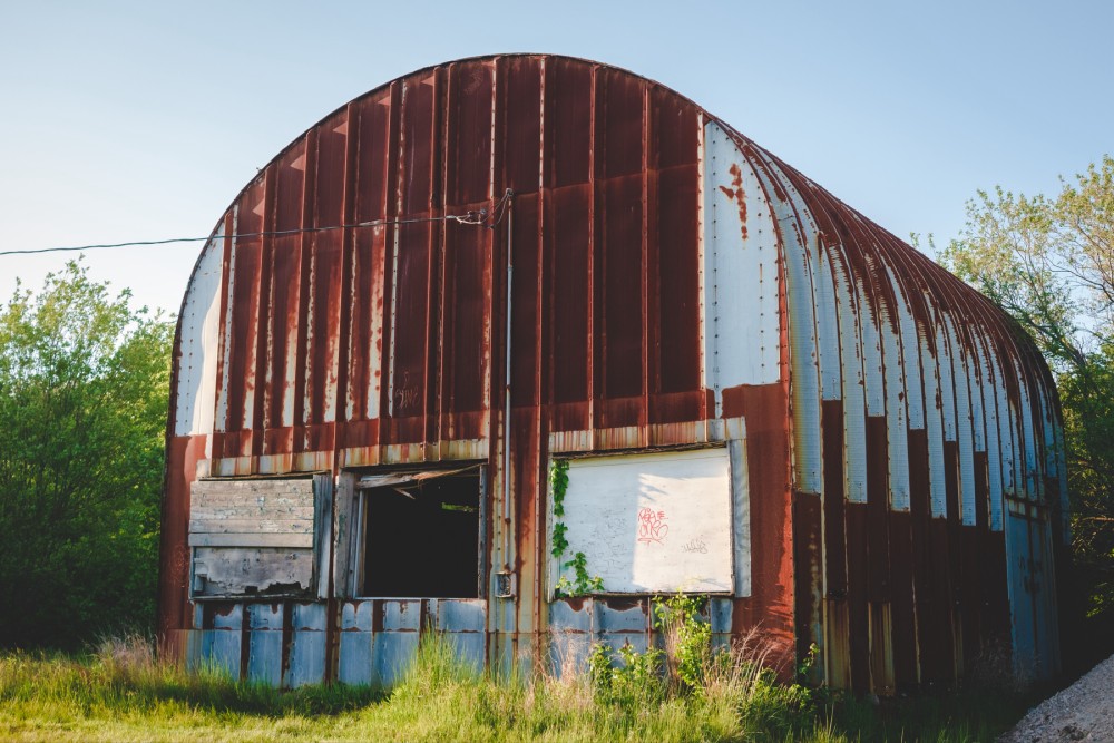 Rusty corrugated metal Quonset hut with open and damaged door, surrounded by wild grass and trees under a clear sky.