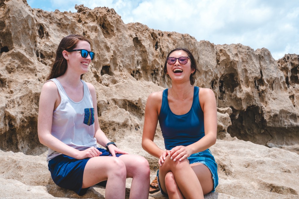 Two women sitting and laughing together on a rocky outcrop, one wearing sunglasses, on a sunny day.