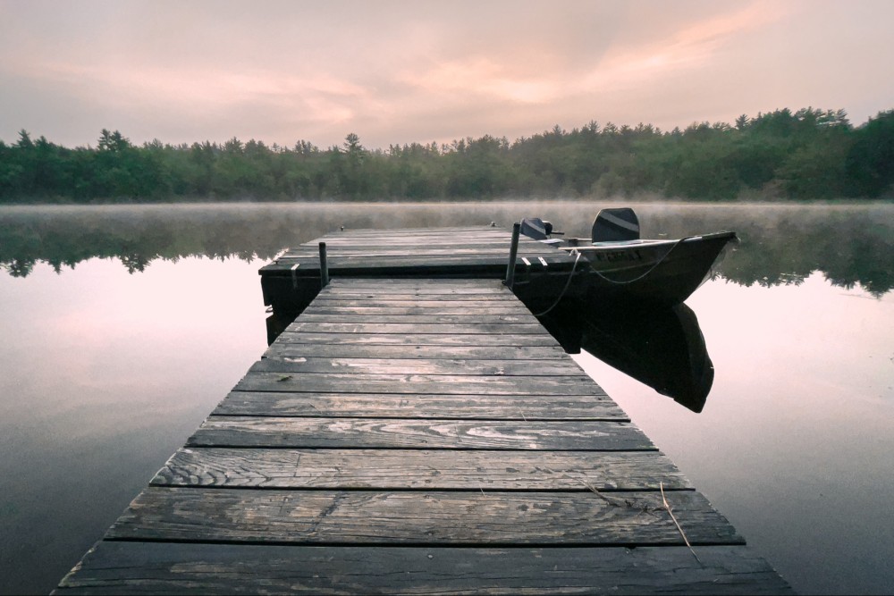 A serene scene of a wooden dock leading out to a glassy lake with a canoe attached to the side. There's a mist over the water and trees reflected on the surface and a pale pink sunrise in the sky