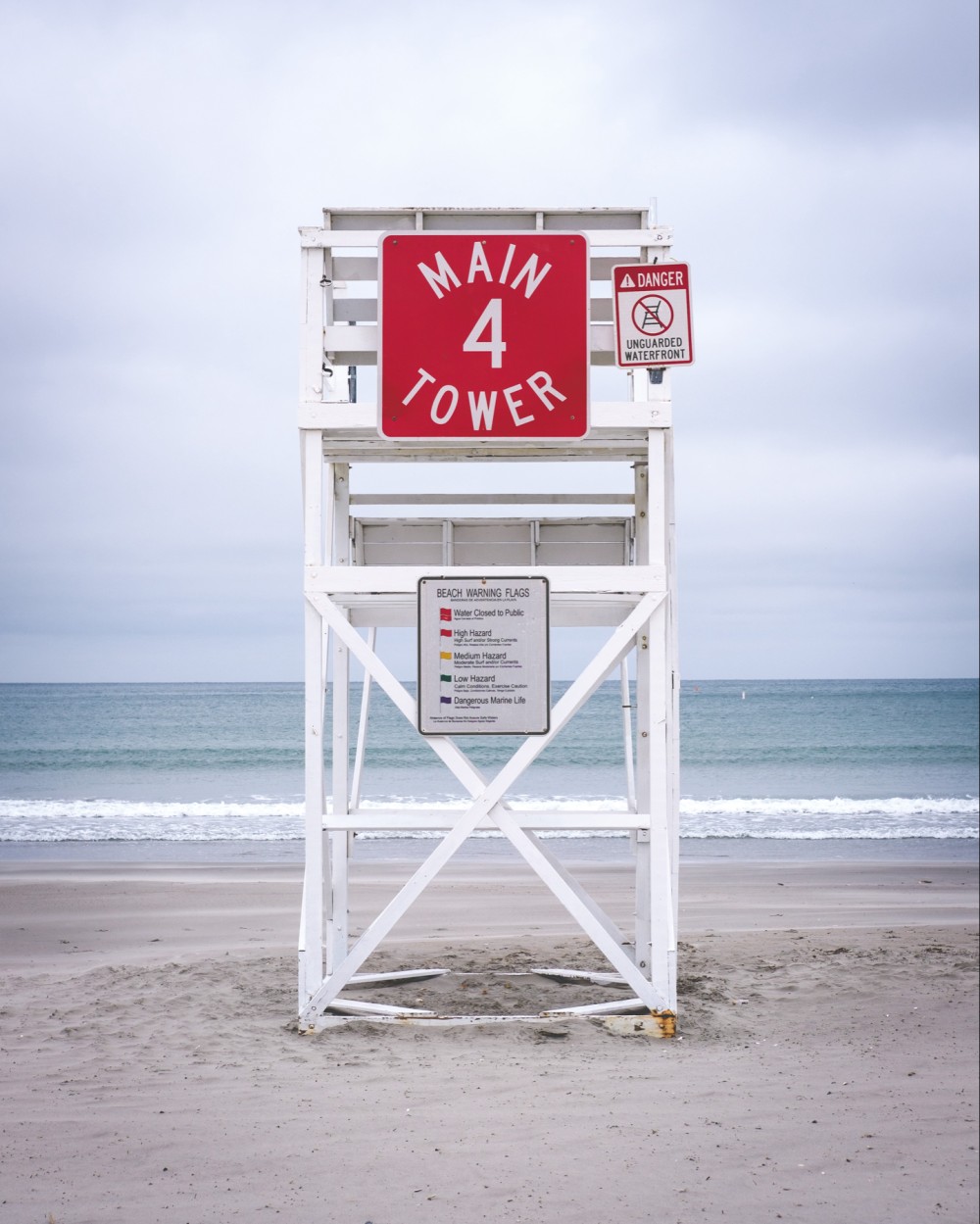 A lifeguard tower labeled "MAIN 4 TOWER" on a beach with a sign next to it indicating "DANGER UNGUARDED WATERFRONT" and a board describing beach warning flags indicating various hazards. The ocean and overcast