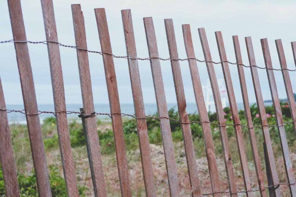 A wooden slat fence with rusty wire along a coastal area with wild vegetation and a blurry lifeguard tower in the background.
