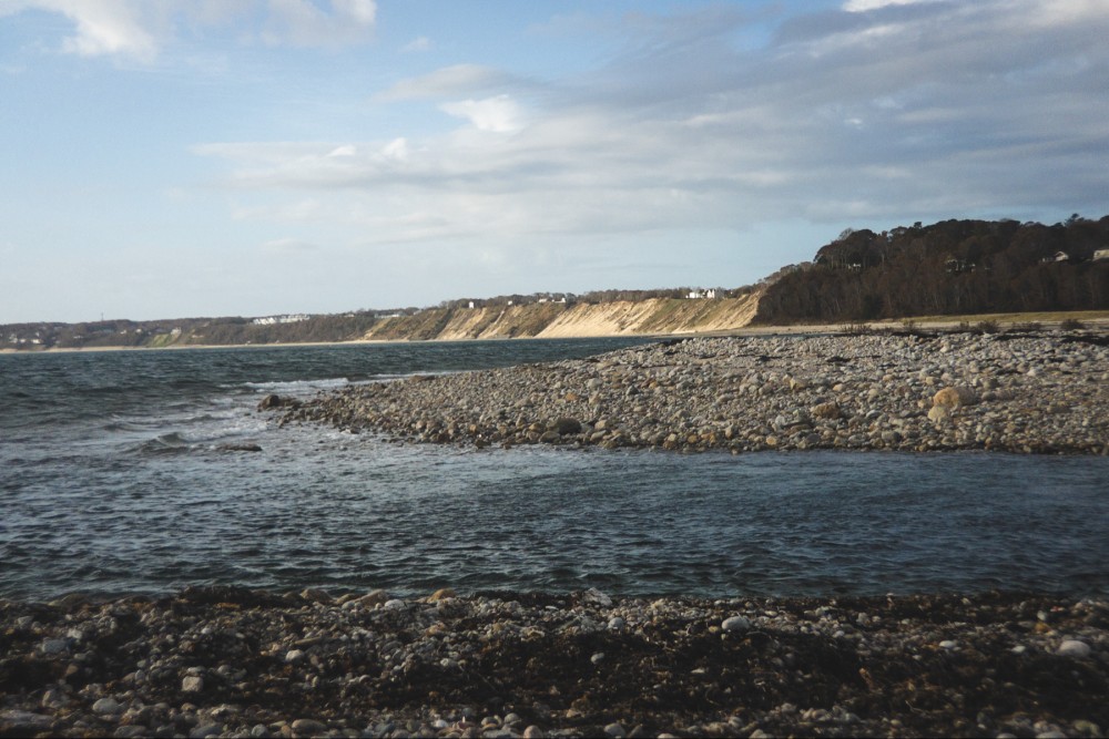 A rocky shoreline with a pebble-strewn beach leading into calm waters, with a bluff in the background featuring sparse vegetation and a few visible dwellings under a partly cloudy sky.
