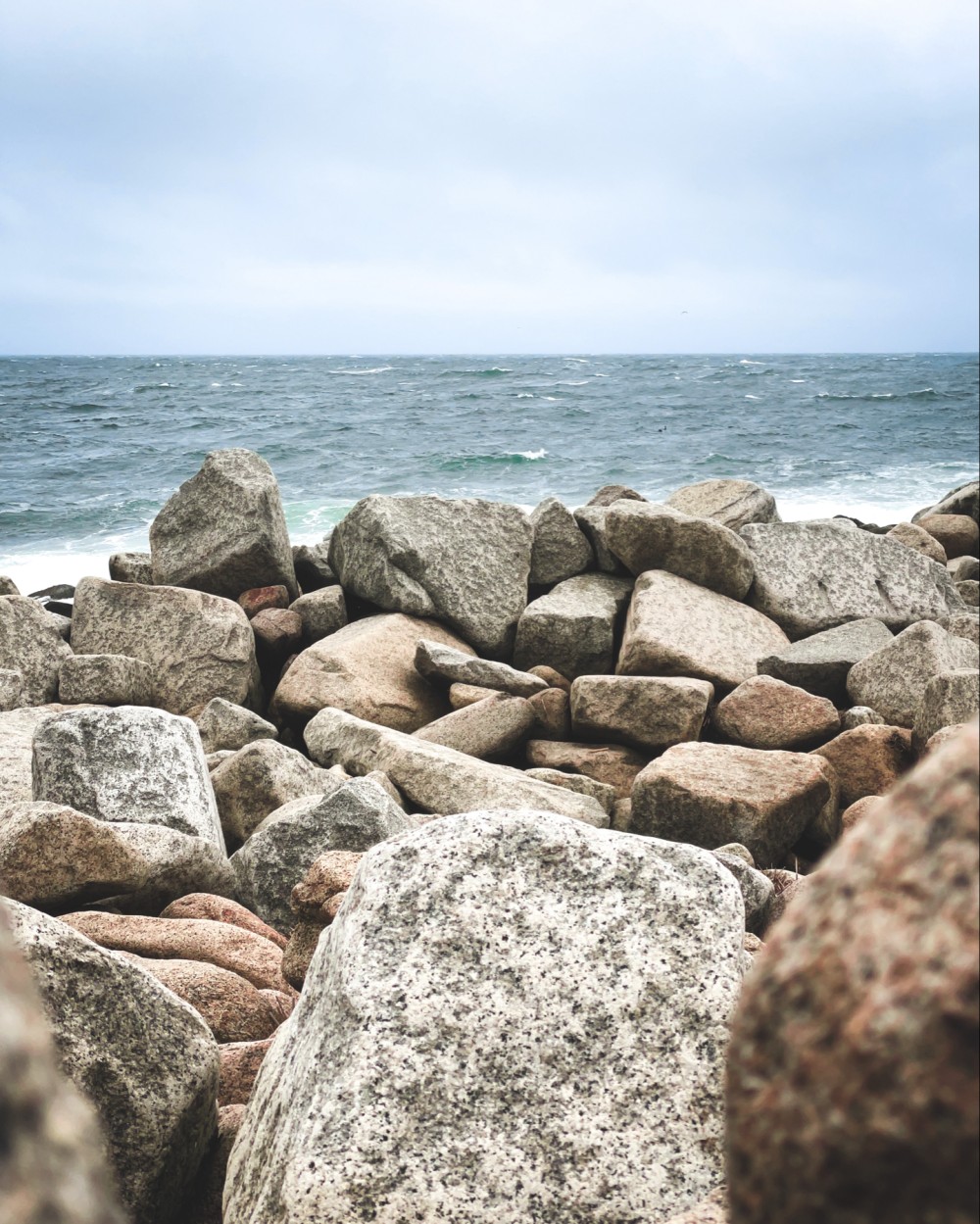 A rocky shoreline with large, multi-colored stones against a backdrop of a choppy sea under a cloudy sky.