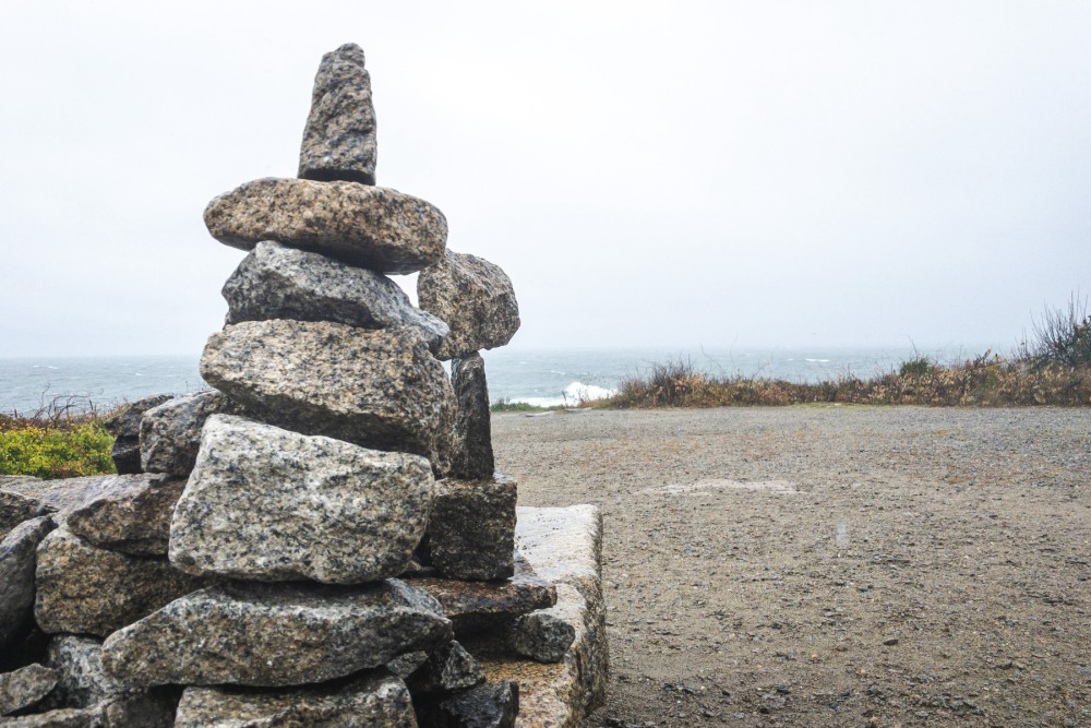 A stone cairn on a sandy surface with the ocean and overcast sky in the background.