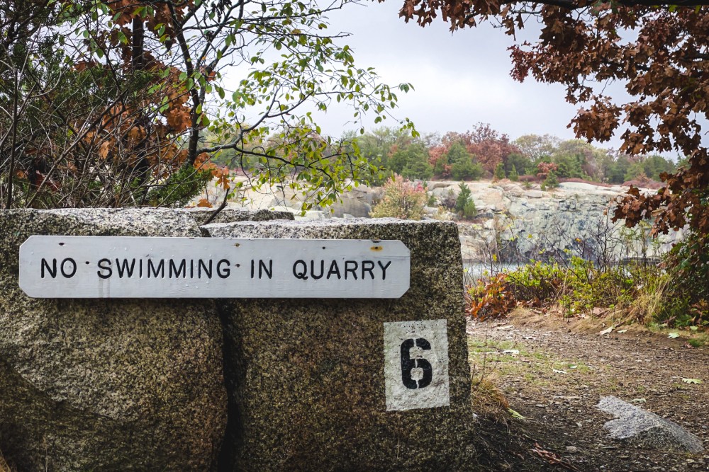 A sign saying "NO SWIMMING IN QUARRY" attached to a large rock with the number "6" painted below it, foliage in autumn colors, and a quarry visible in the background.