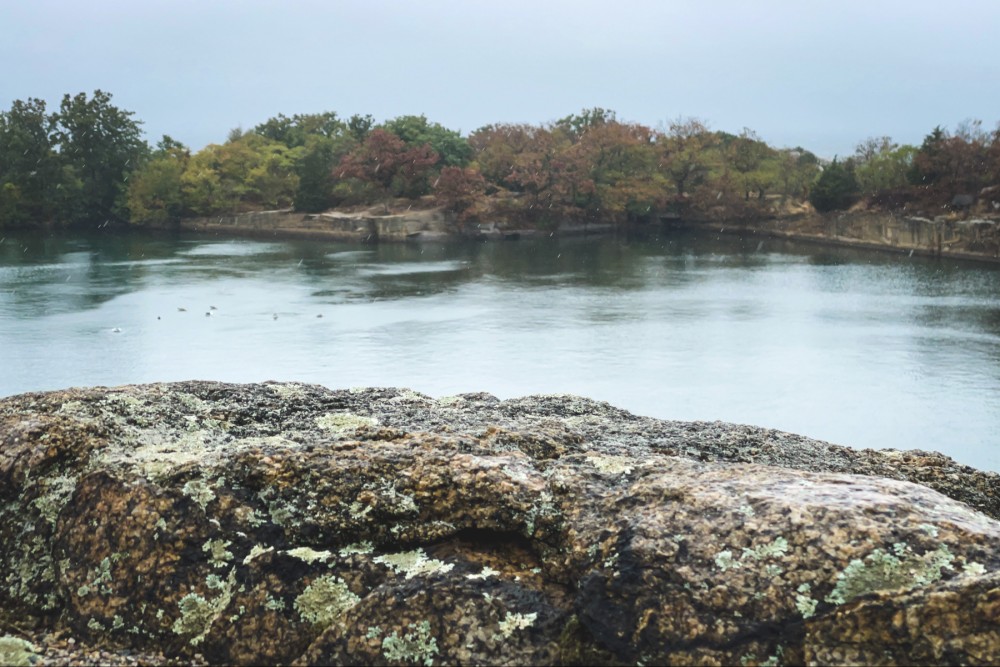A tranquil quarry with a rocky foreground covered in lichen and autumn-colored trees in the distance.
