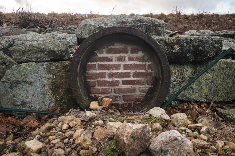 A circular opening set in a stone wall, sealed with bricks, surrounded by scattered rocks and some foliage.