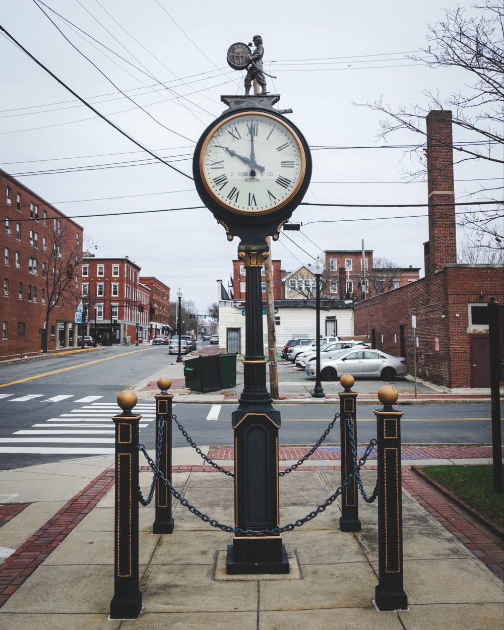 A street clock on a sidewalk with a decorative statue on top, surrounded by chains connected to posts, with industrial brick buildings and parked cars in the background.