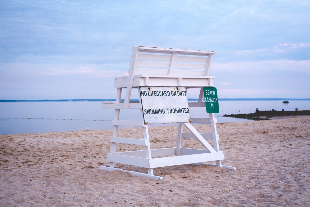 A lifeguard tower on an empty beach with signs stating "No Lifeguard on Duty" and "Swimming Prohibited," along with a sign showing "Beach Capacity 75” at sunrise