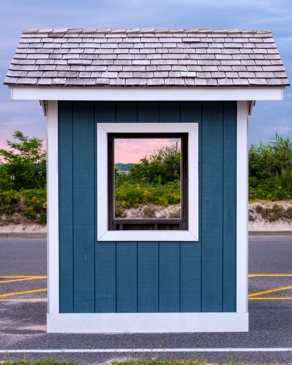 A small shelter with a shingled roof and teal walls by the side of a parking lot, featuring a window that frames a view of a colorful beach sunrise sky.