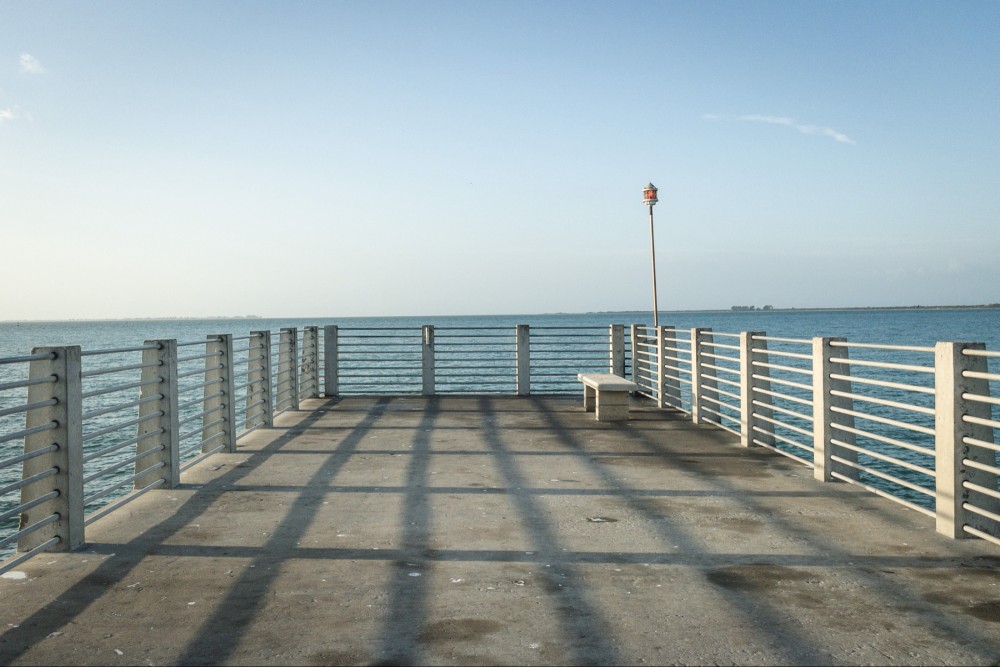 A pier with a grid of shadows extending into the ocean with a bench, railings, and a light pole against a backdrop of a clear sky and distant land.