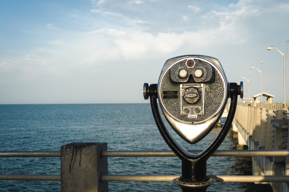 A tower viewer binoculars overlooking the sea from a pier with a clear sky.