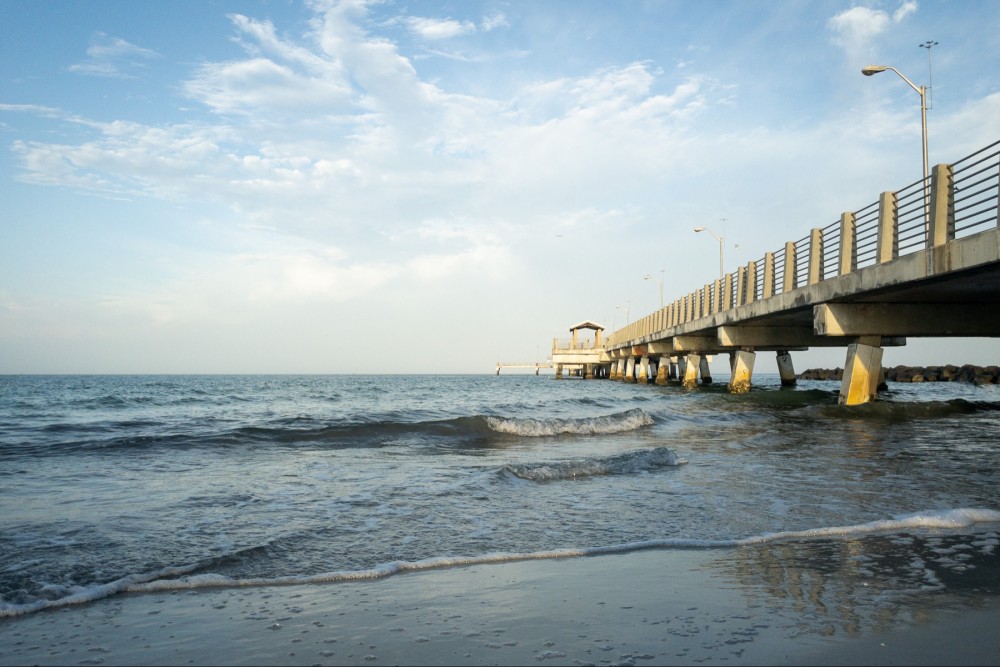 A long pier extending into the ocean with a small shelter at the end, gentle waves on the shore, and a blue sky with wispy clouds.