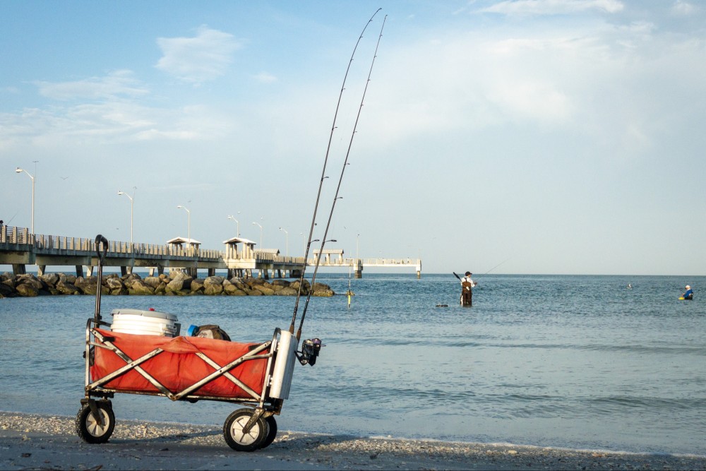 A fishing cart with gear on a beach, with people fishing in the water and a pier in the background.