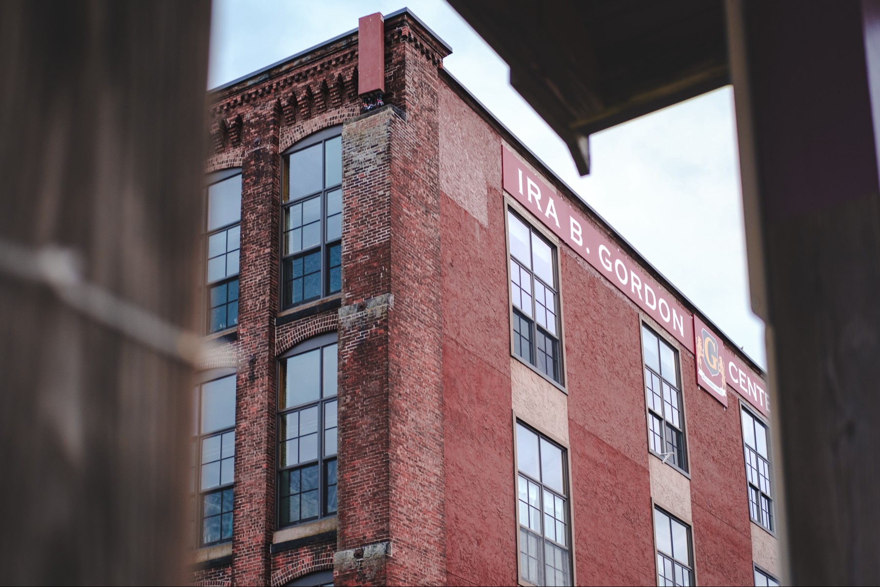 An upward shot of an industrial brick building with large windows and a sign that reads "IRA B. GORDON CENTER” framed through a wooden structure