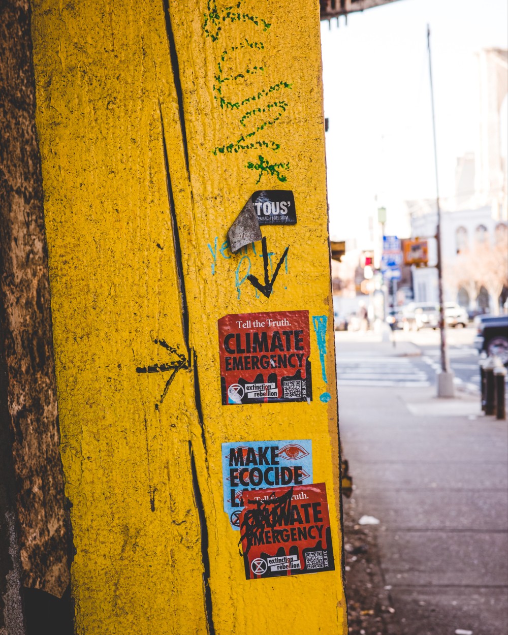 A weathered yellow wall with cracks, hand drawn symbols, and various stickers along a sidewalk in an urban area