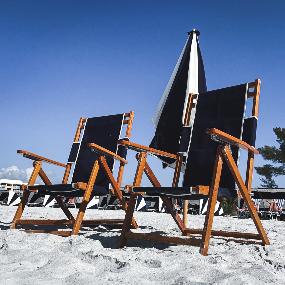 Photo of two navy blue beach chairs with warm wooden frames in front of a blue and white umbrella on a beach with a completely blue sky