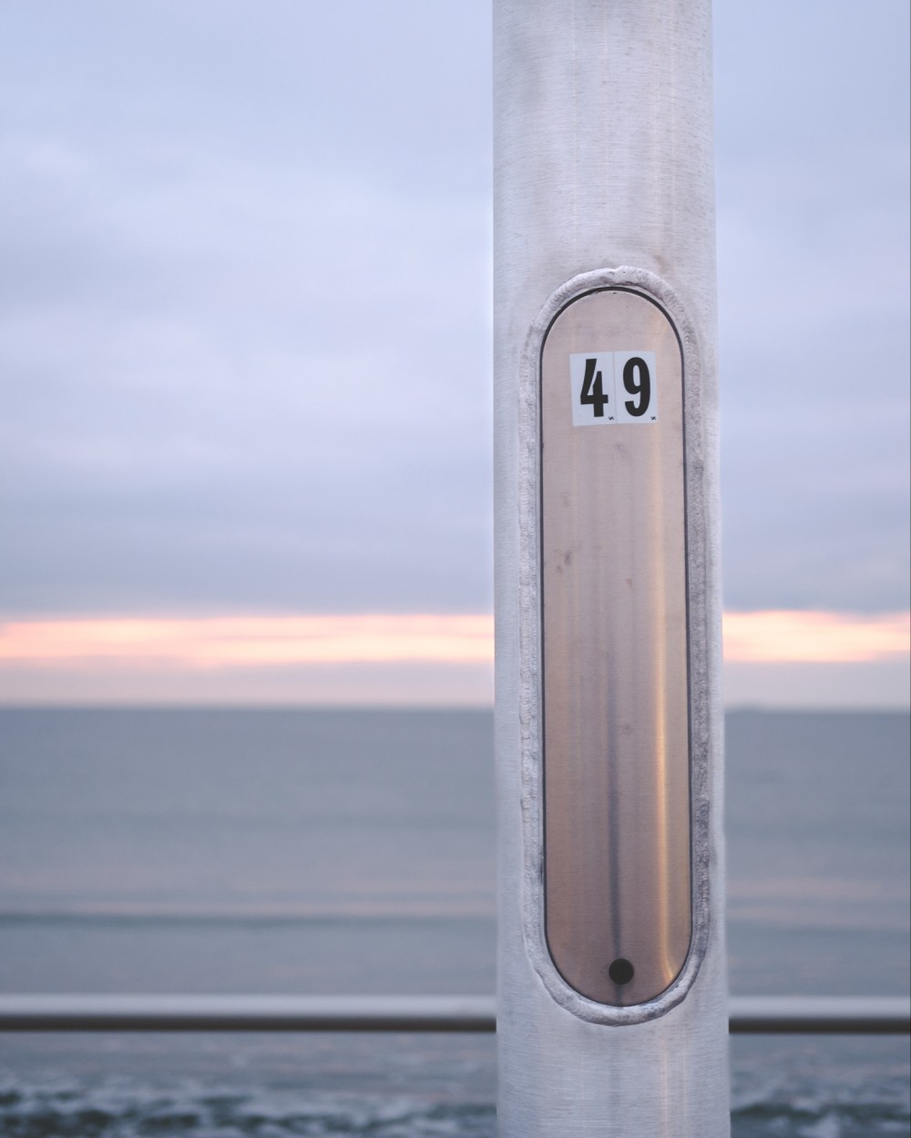 A close-up of a metallic pole with a number plaque reading "49" against a blurred background of a pale sunrise over the sea.