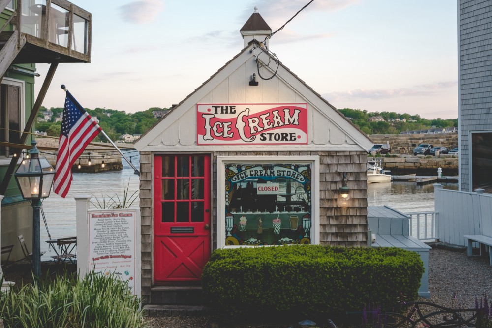 A quaint ice cream store with a red door and an American flag, situated by a waterfront with boats and parked cars in the background, under a sunrise sky. The store's sign reads "CLOSED."