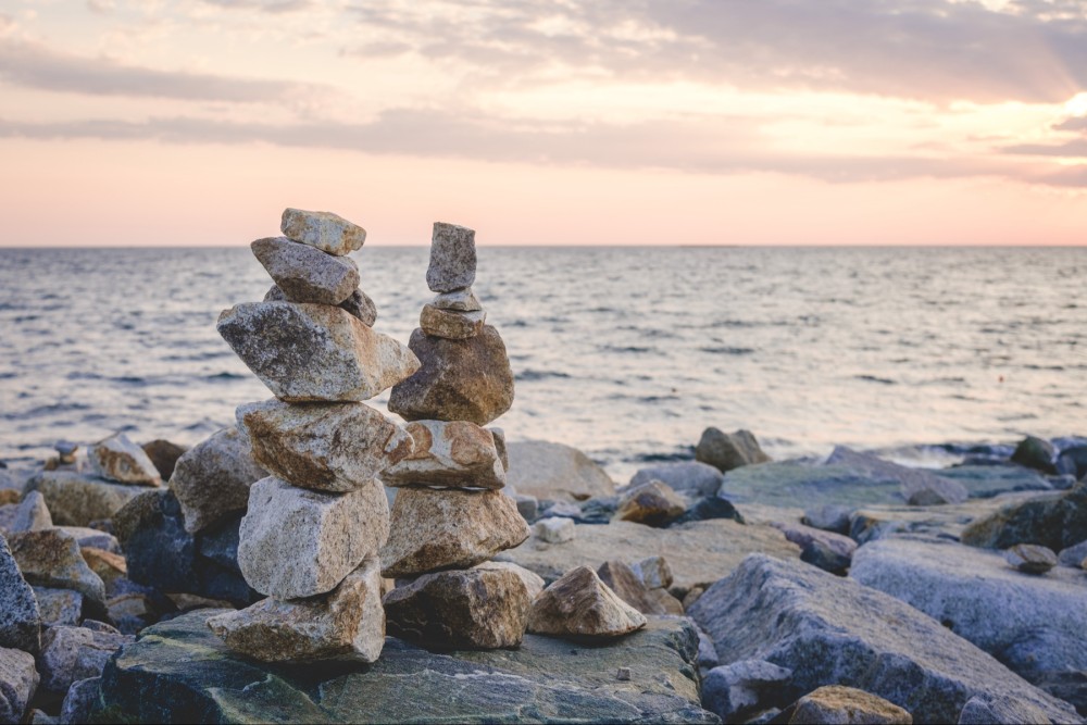 Stacked rock cairns on a rocky shoreline with the ocean in the background during sunrise.