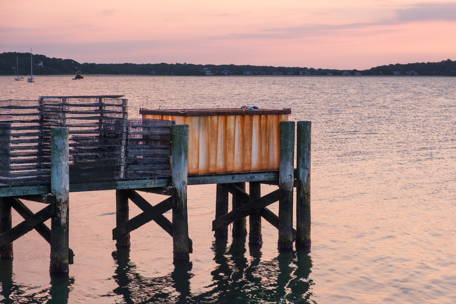 A serene waterside scene at sunrise featuring a weathered wooden dock with rusty, metallic containers and stacks of fishing traps. In the background, calm waters reflect the soft hues of the sky, with a distant shoreline and a few anchored sailboats.