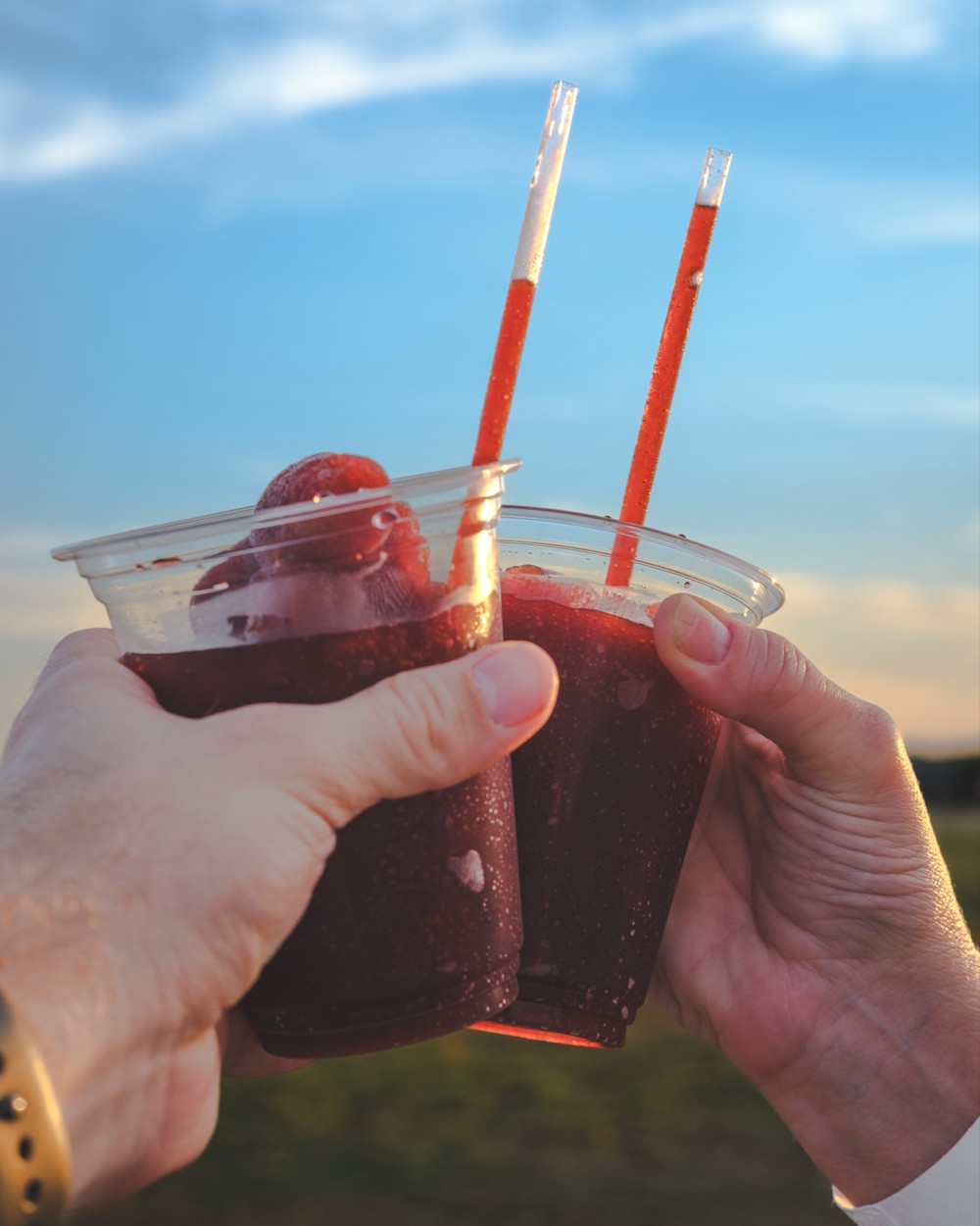 Two hands holding clear plastic cups with a red-colored frozen drink and straws against a blue sky background.