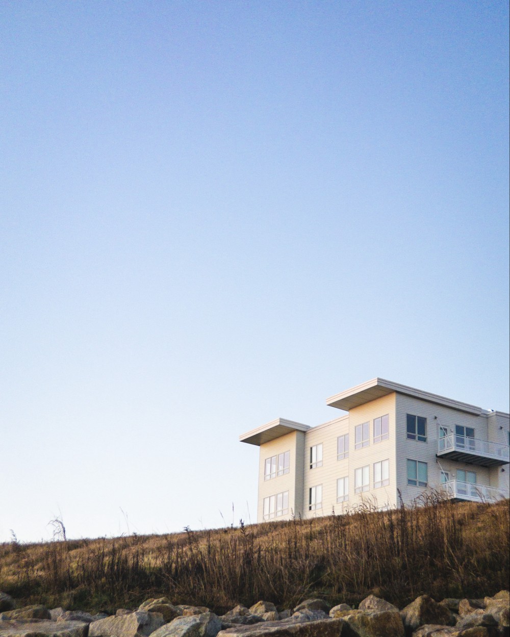 A modern, multistory residential building on top of a grassy hill with a clear blue sky in the background and a rocky foreground.
