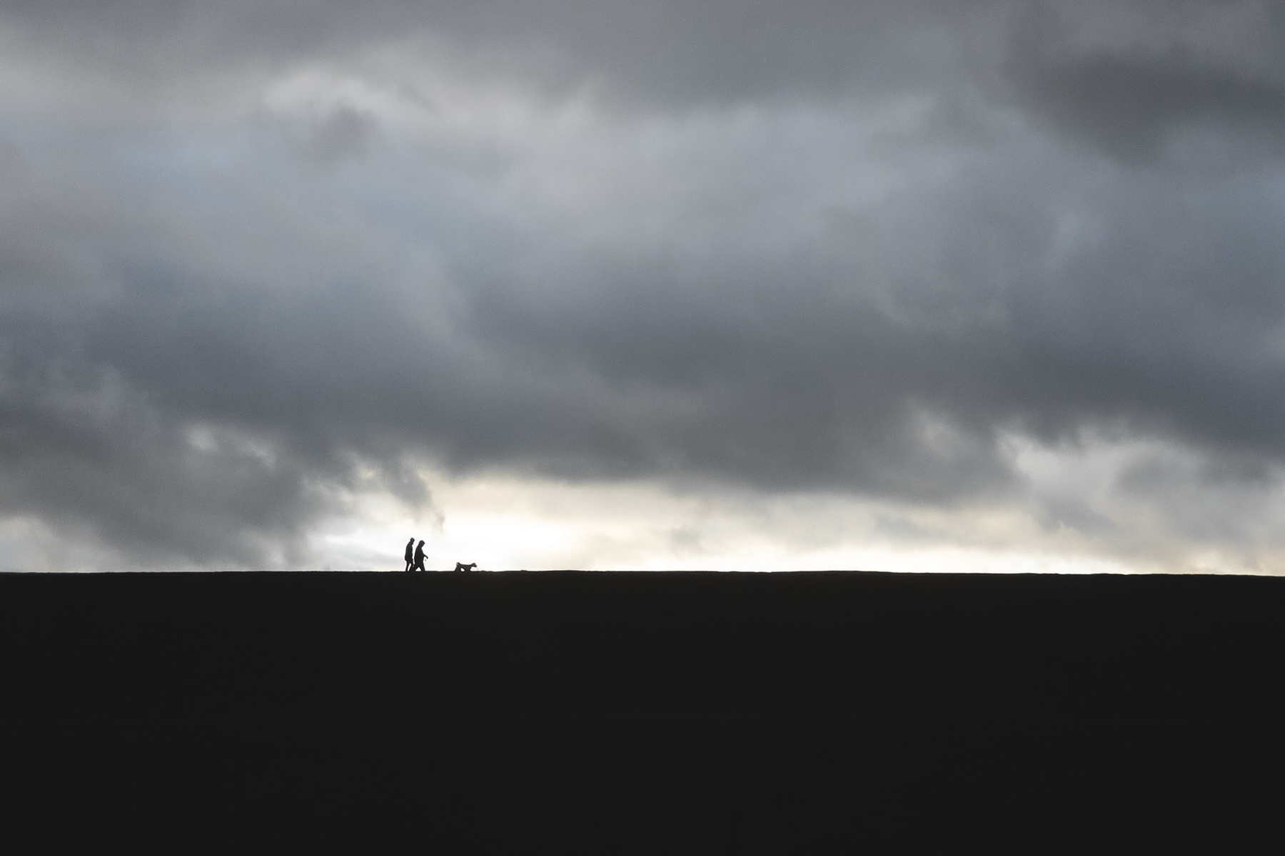 Silhouetted figures walking a dog on a ridge under a dramatic cloudy sky