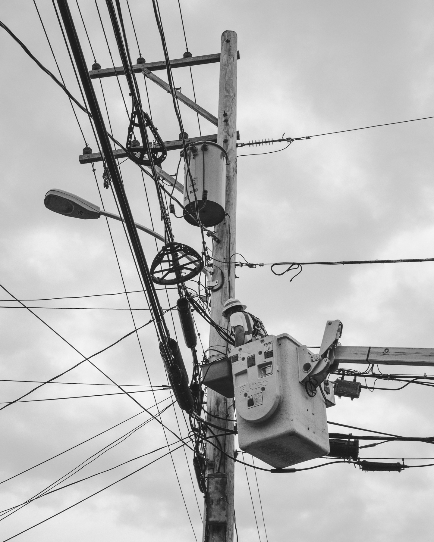 A black and white photo of a utility pole with a transformer, multiple cables, and a street light against a cloudy sky.