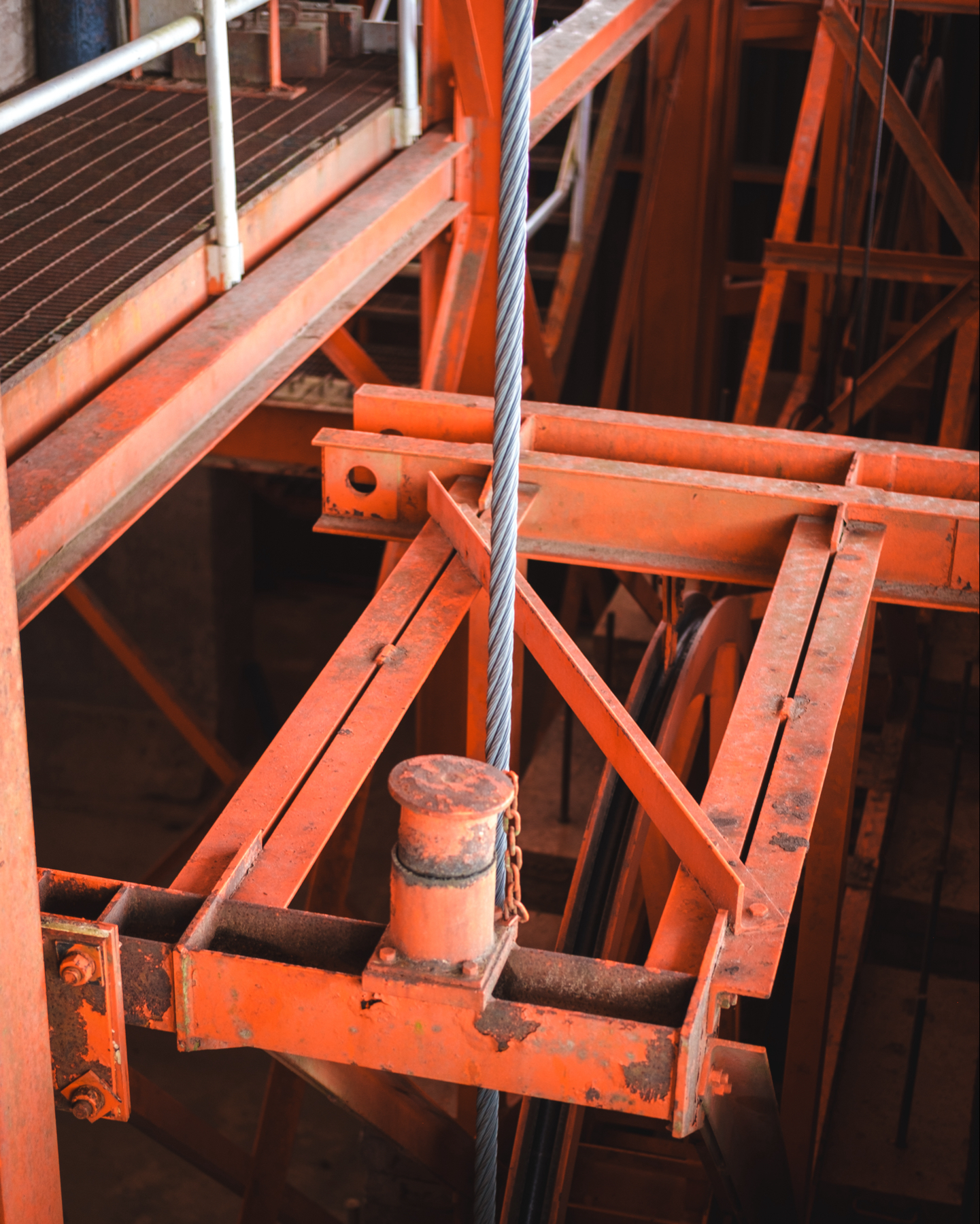 Industrial crane pulley with a thick steel cable, set against a backdrop of orange metal construction with visible rust and wear.