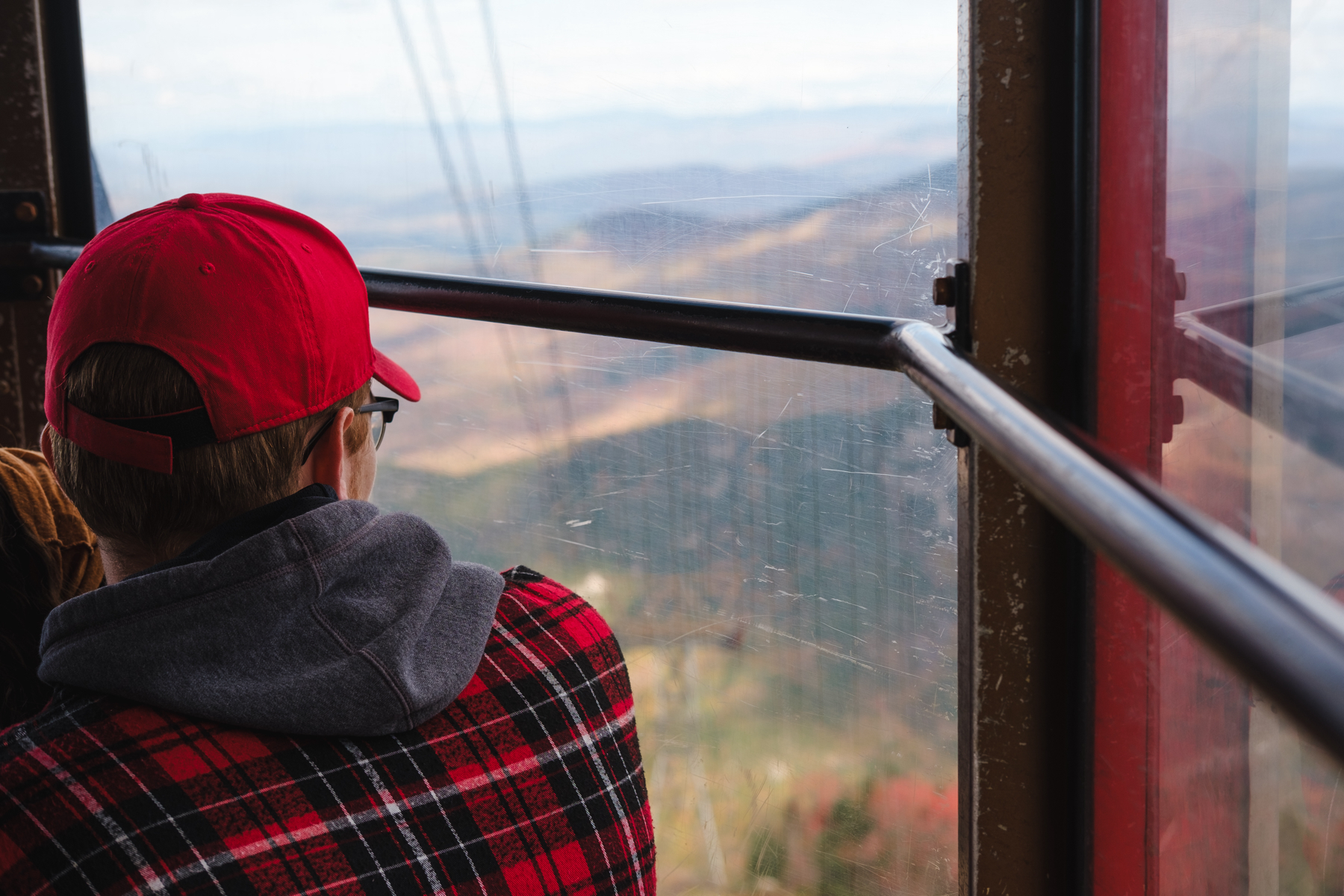 A person wearing a red baseball cap, a red flannel, and glasses looking out the window of a cable car with a scenic mountain landscape in the background.