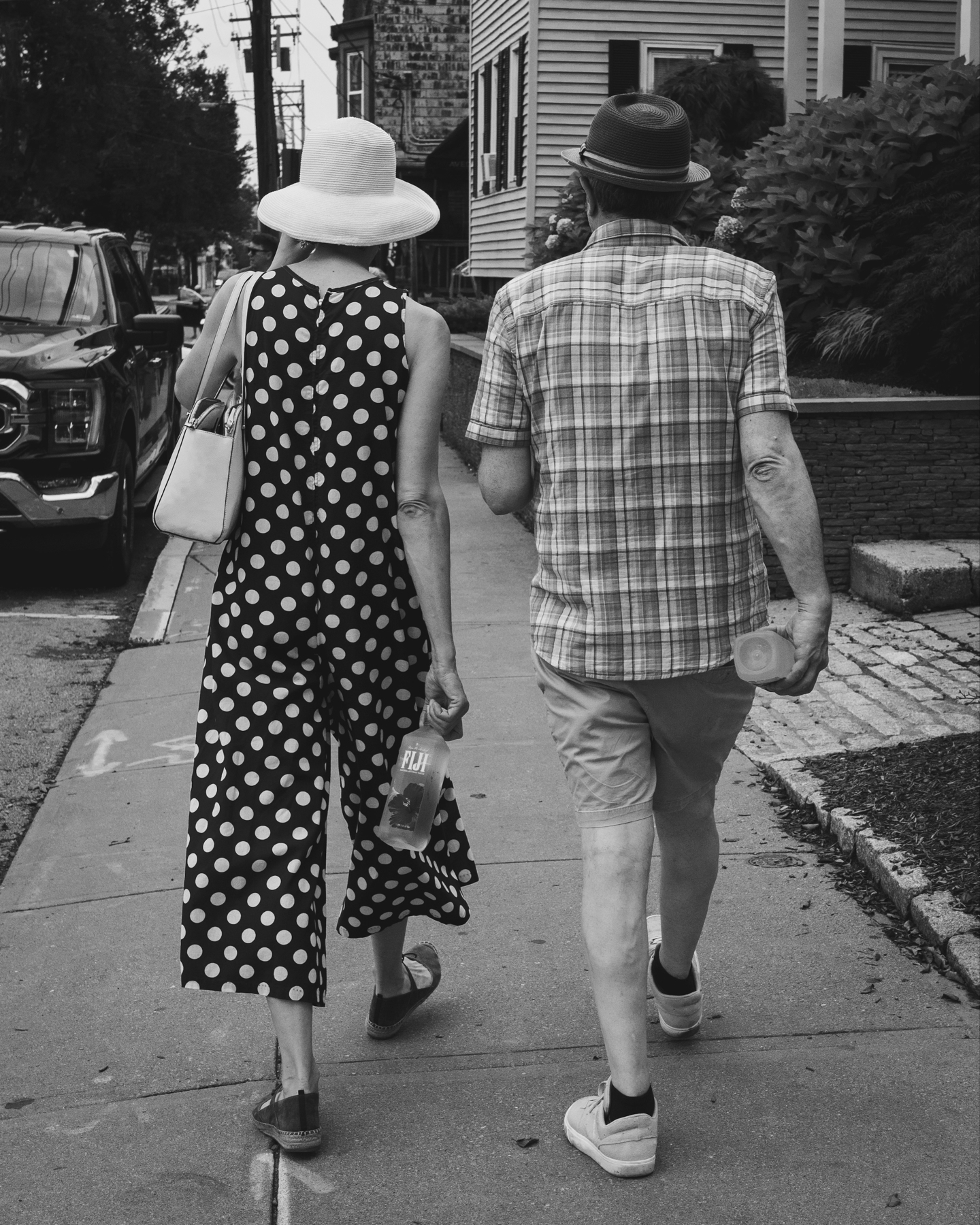 A black and white photo of a two people walking side by side on a sidewalk. One is wearing a polka dot dress and a white sun hat, carrying a white handbag and a water bottle. The other is in a plaid shirt and dark brimmed hat, also carrying a water bottle.