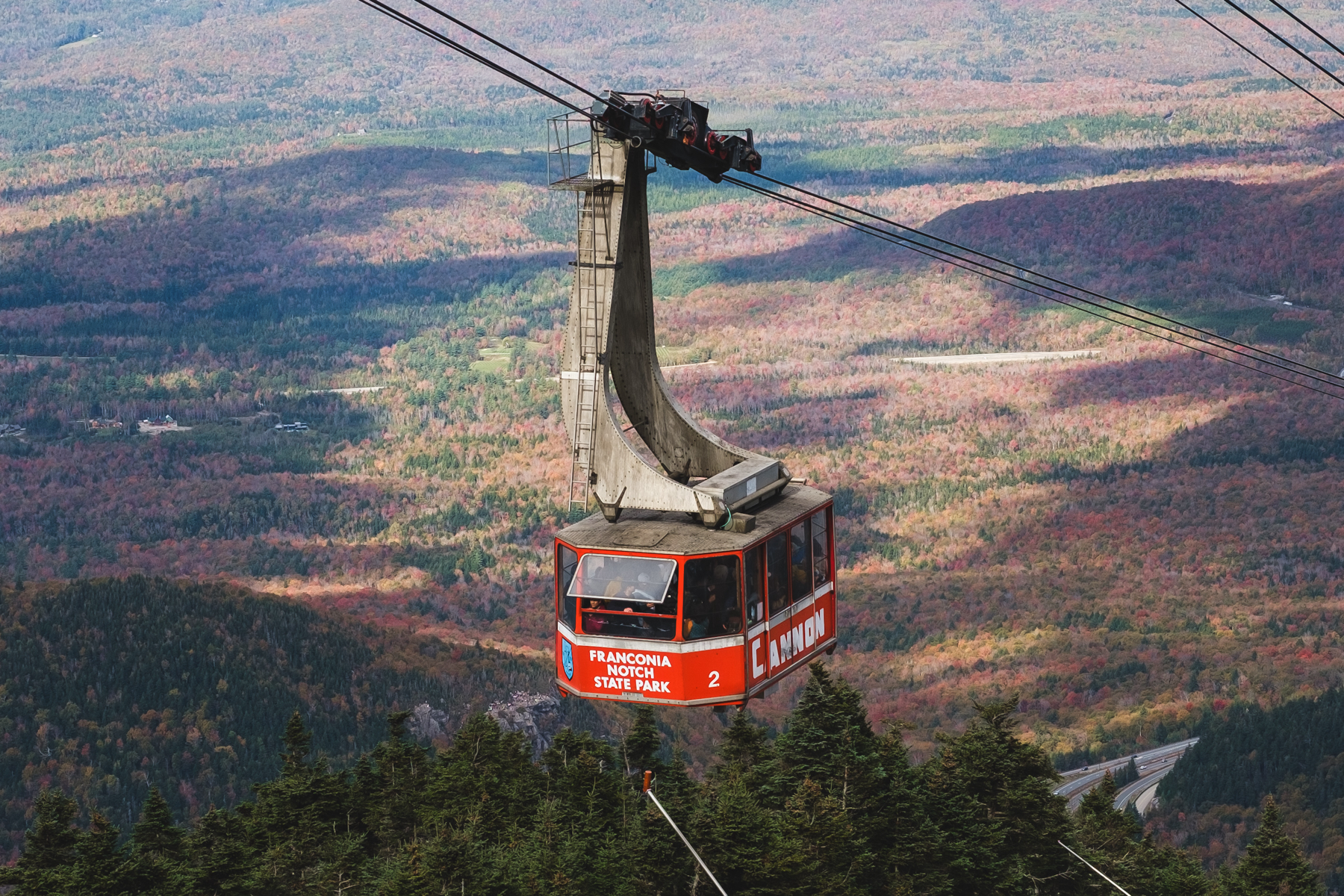 A red cable car with “Franconia Notch State Park” and “Cannon” written on it above mountains with autumn foliage.