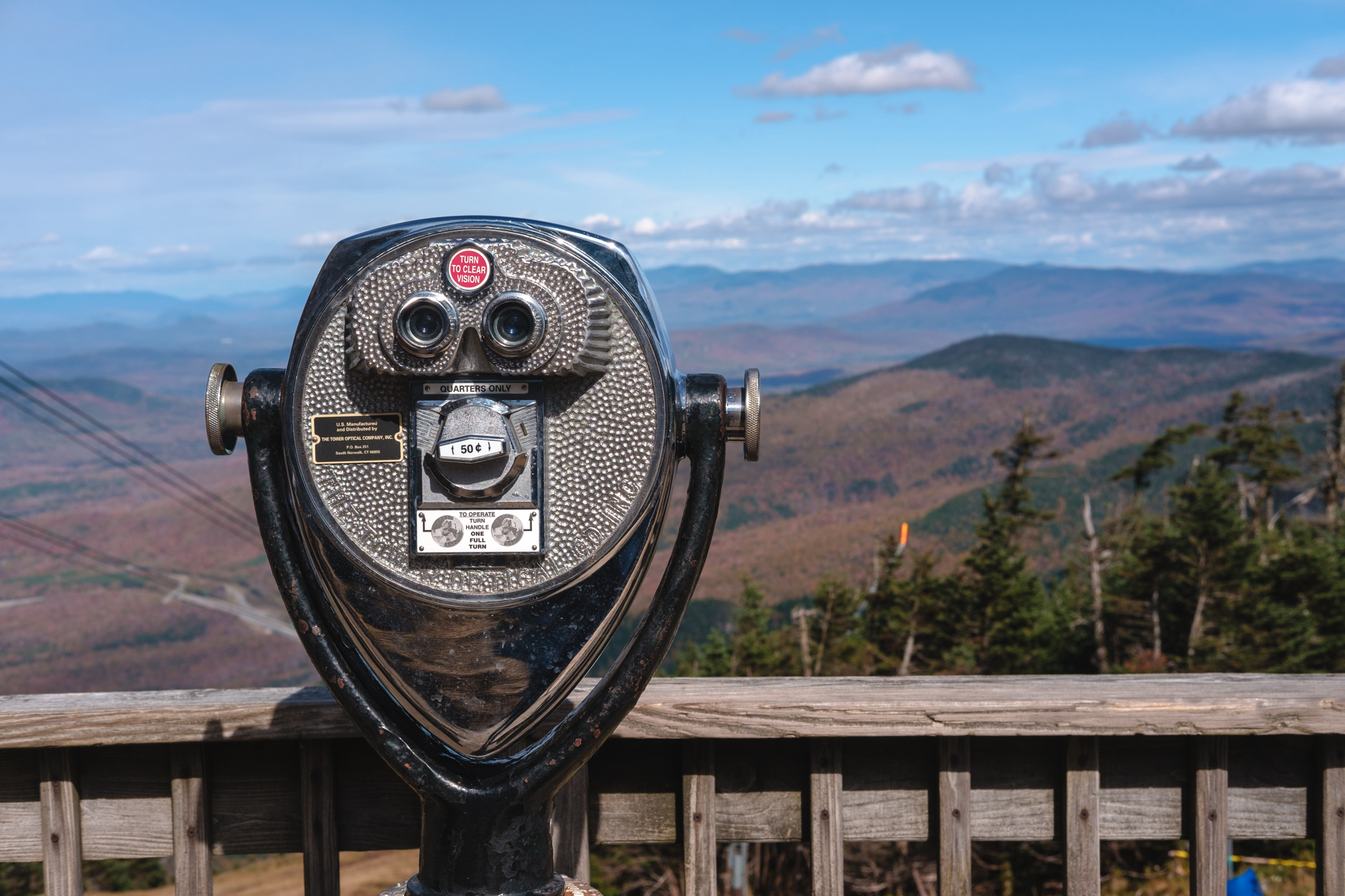 Tower viewer coin-operated binoculars on an overlook deck with expansive mountainous landscape in the background under a blue sky with clouds.