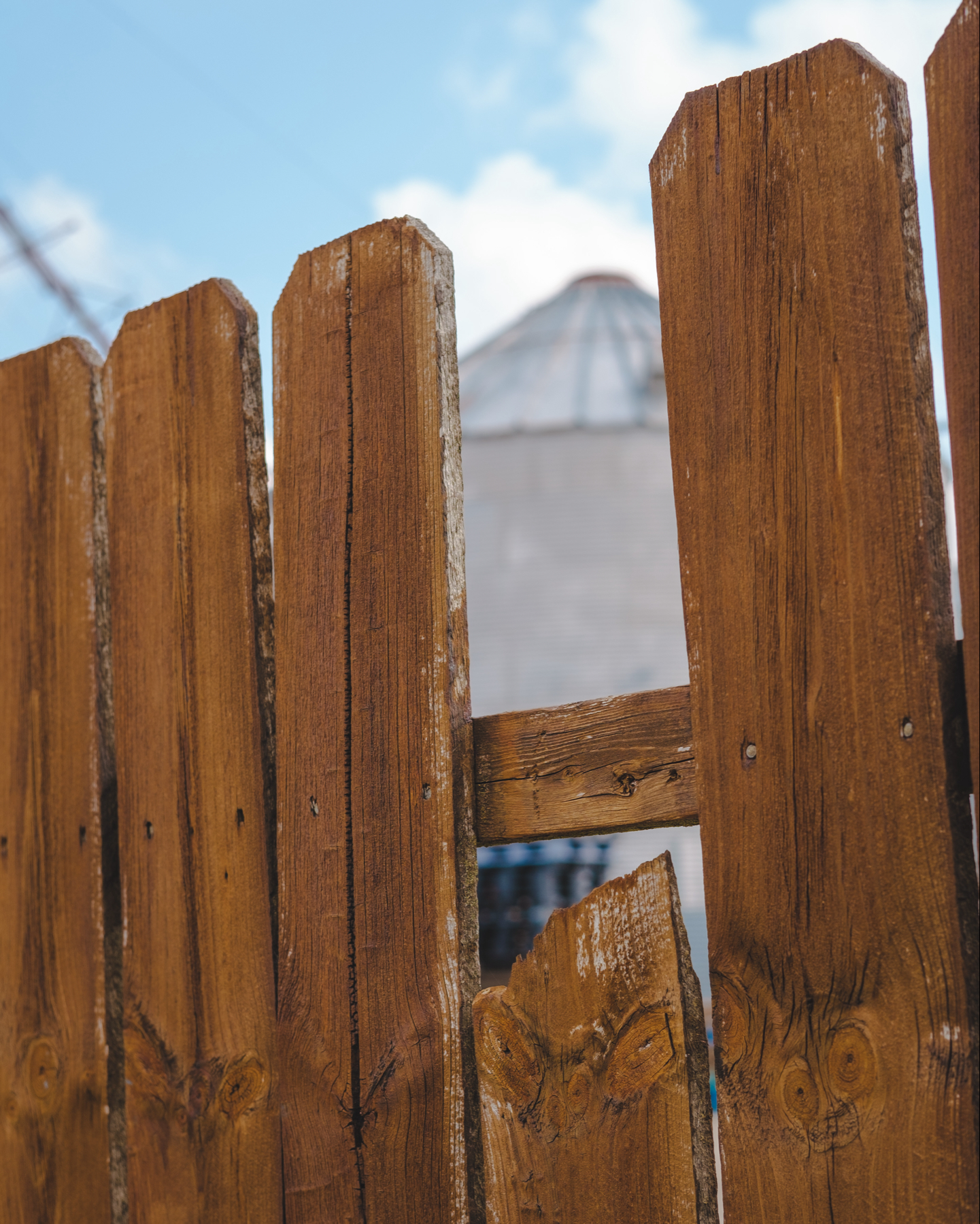 Wooden fence with a broken plank framing a grain silo and blue sky 