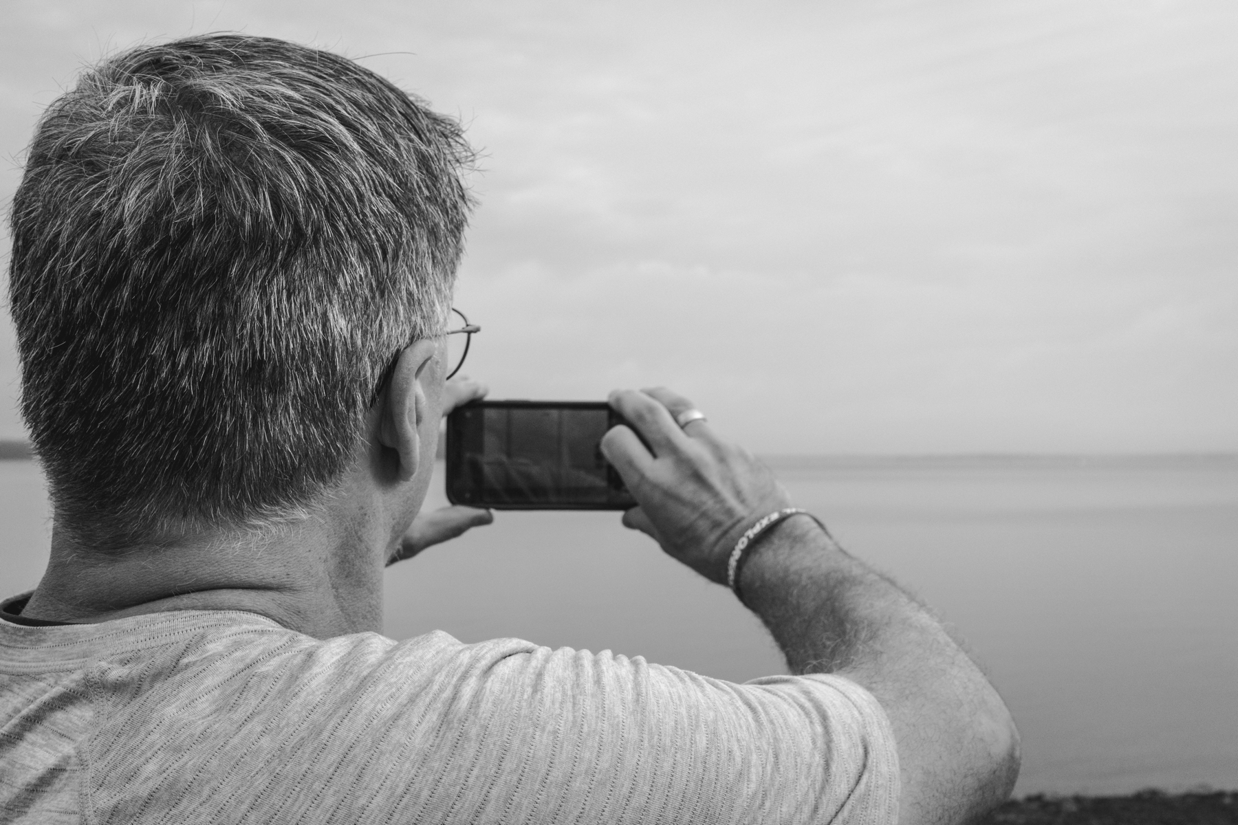 A black and white photo over the shoulder of a man taking a smart phone picture of the ocean