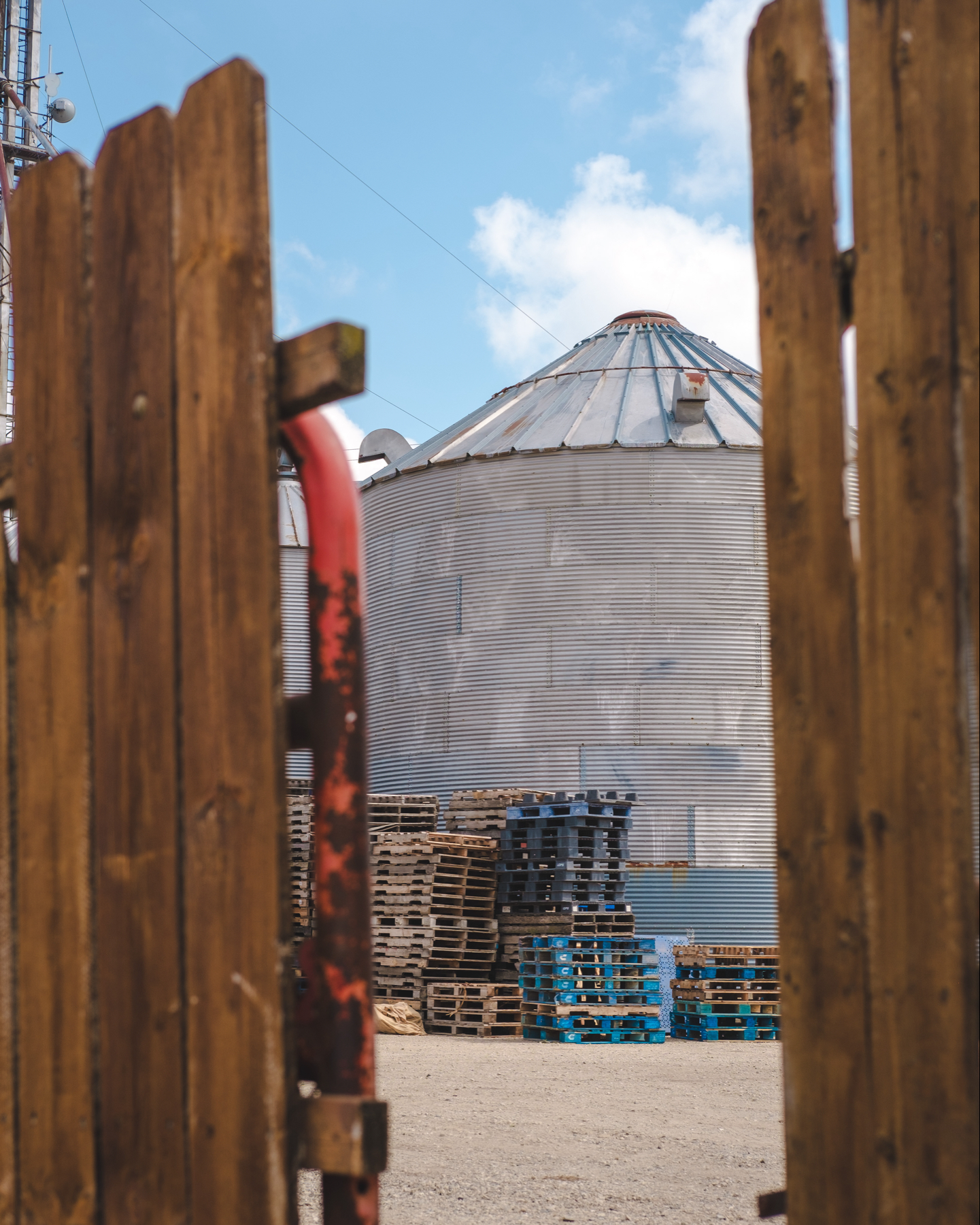 A grain silo seen between the slats of a warm brown wooden fence, with stacks of wooden and blue plastic pallets in the background.