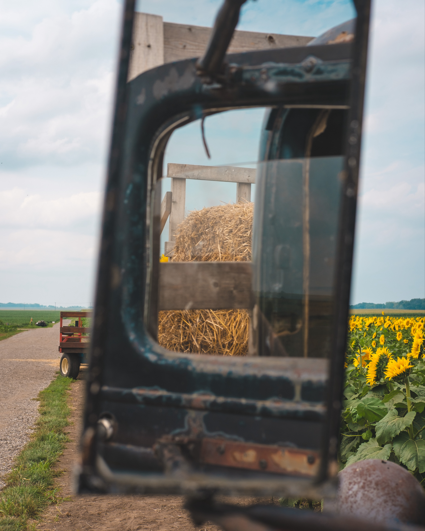 A rustic scene framed through the mirror of an old tractor cab, showing a hay bale on a trailer with a sunflower field in the background.