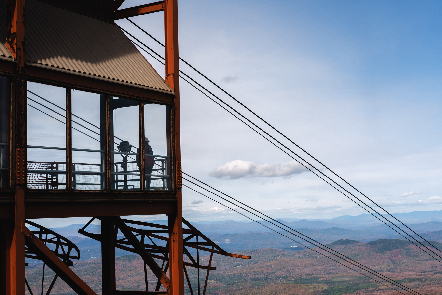 A ski lift tower with a lone person standing on the observation deck, thick cables running diagonally across the frame, and a panoramic view of a mountainous landscape under a blue sky with clouds.