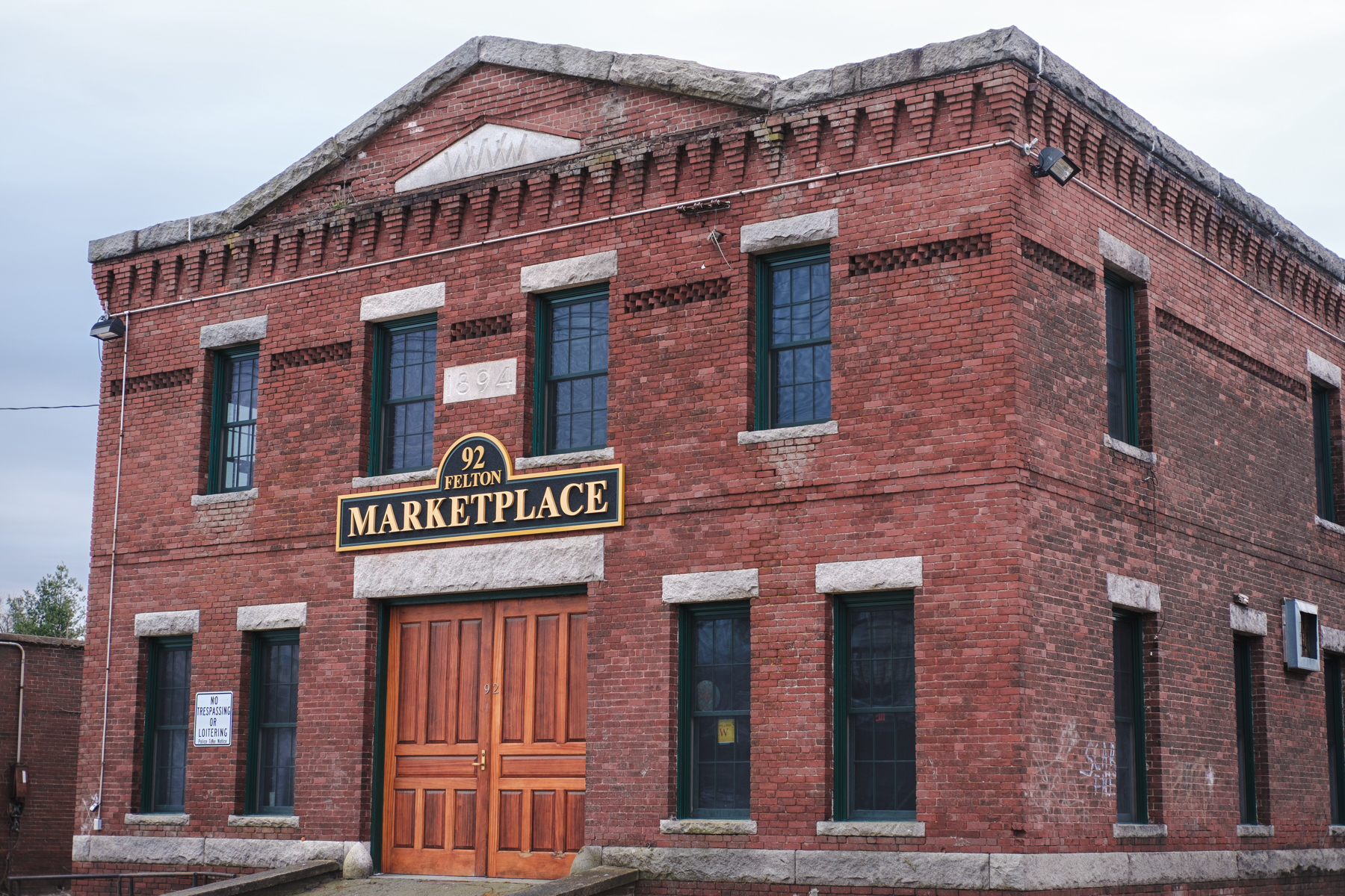 A two-story red brick building with the signage “92 FELTON MARKETPLACE” and the year “1894” displayed near the roof. The building features large wooden double doors, rectangular windows, and decorative brickwork along the edges. 