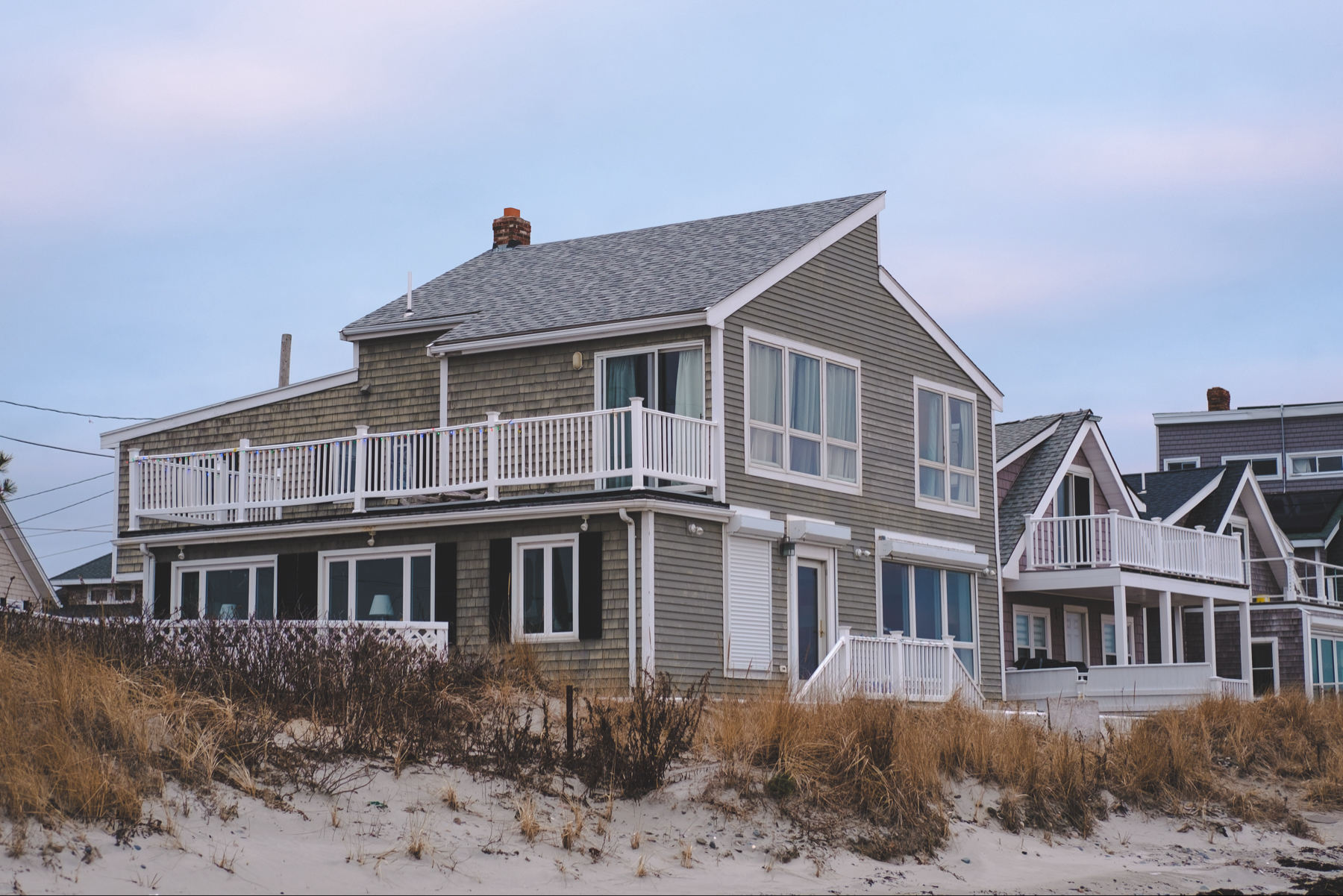 A beachfront house with gray siding and white railings sits on sandy ground with sparse grass. An overcast sky looms above.