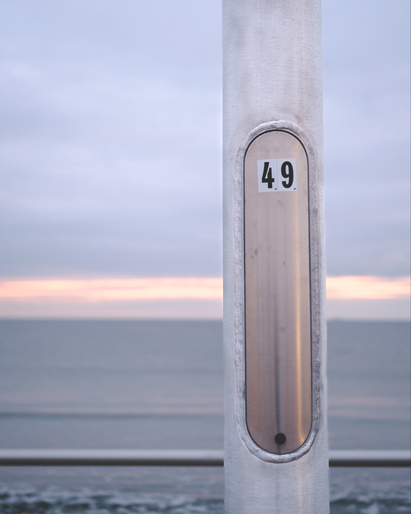 A close-up of a metallic pole with a number plaque reading “49” against a blurred background of a pale sunrise over the sea.
