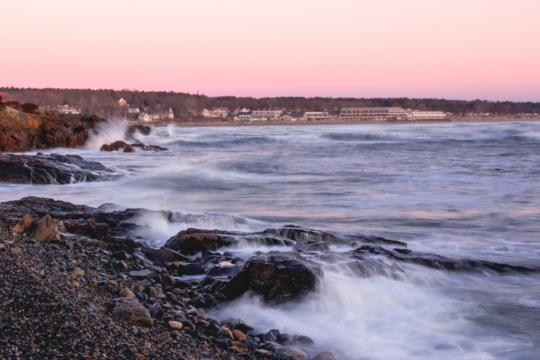 Seascape at sunrise with waves crashing against rocky shoreline, pebbly beach in foreground, and large waterfront buildings in the distance under a pink-hued sky.