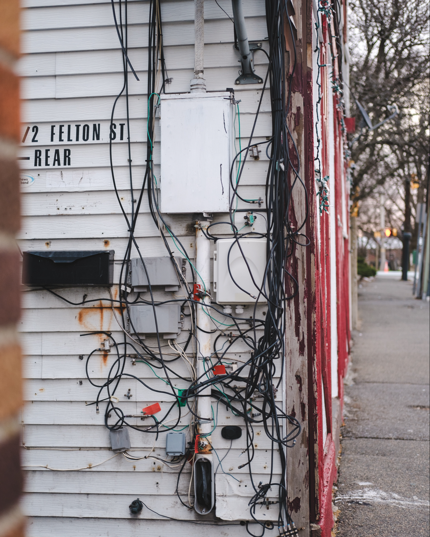 A cluttered array of electrical wires and boxes on the corner of a multi family home. The building shows signs of weathering. A sidewalk passes the front of the building. 