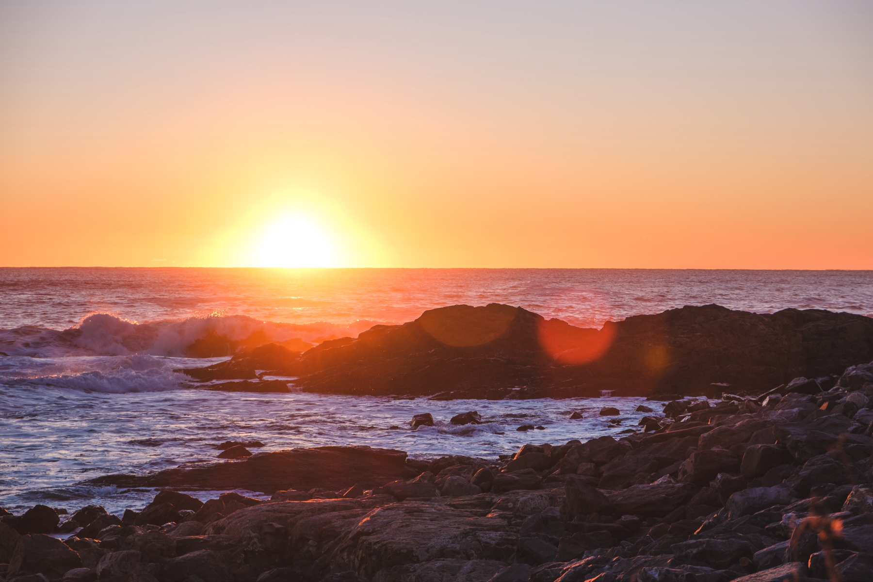 Sunrise over a rocky coastline with waves crashing against the rocks and a warm orange sky.