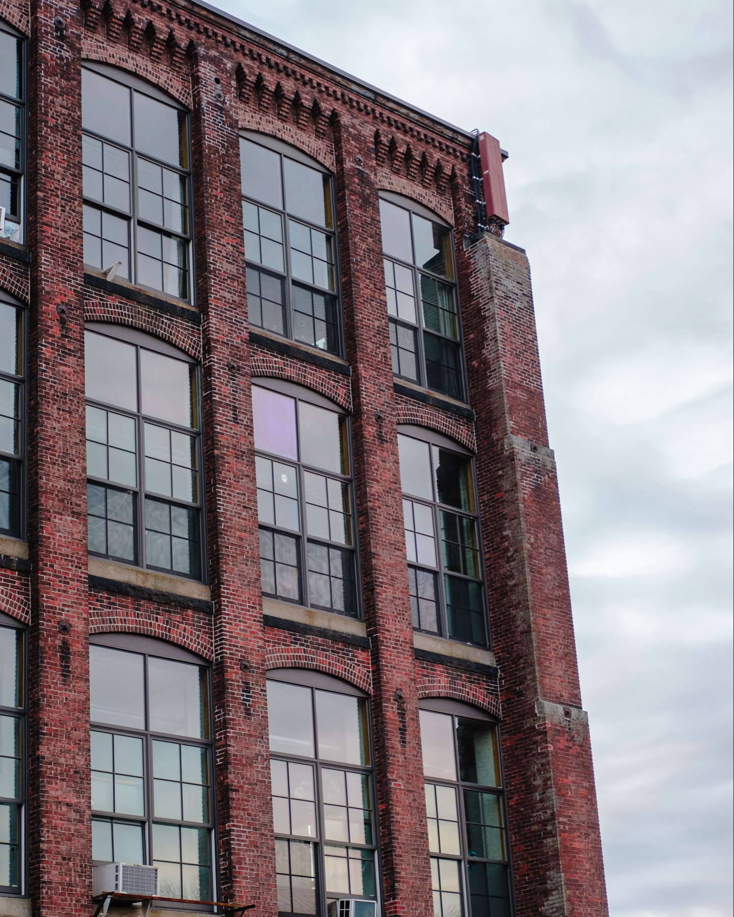 A tall red brick industrial building with large windows against a cloudy sky.