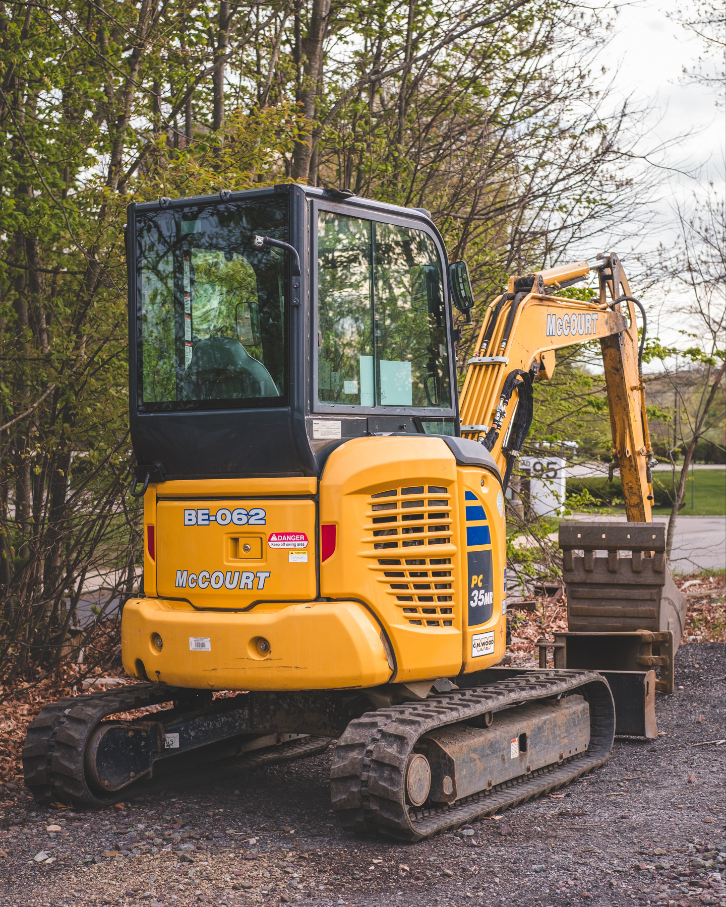 An empty yellow compact excavator with “McCourt” written on the arm, parked on a gravel surface with trees in the background.